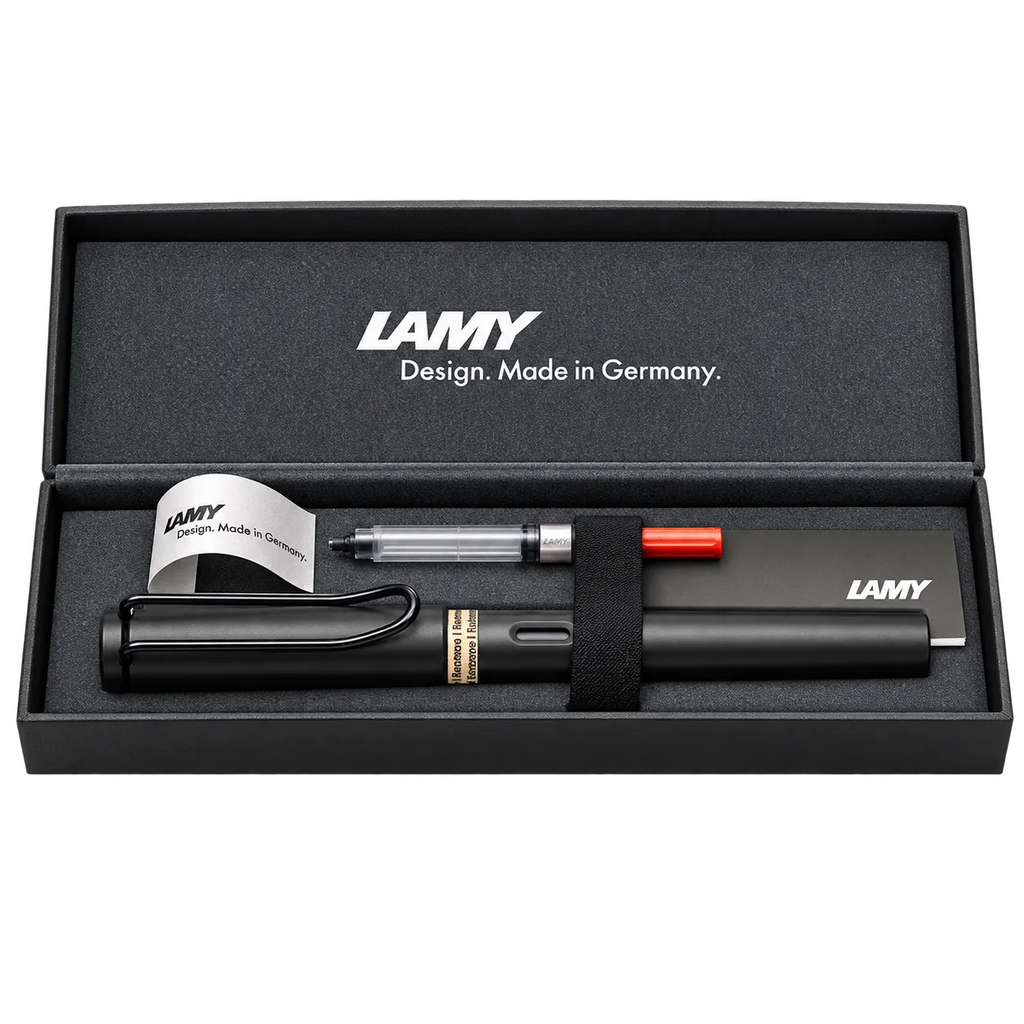 LAMY pen set in an open black presentation box, including a black pen, a clear and silver top-load pen, a small orange pen cap, and branded paper inserts on a white background.
