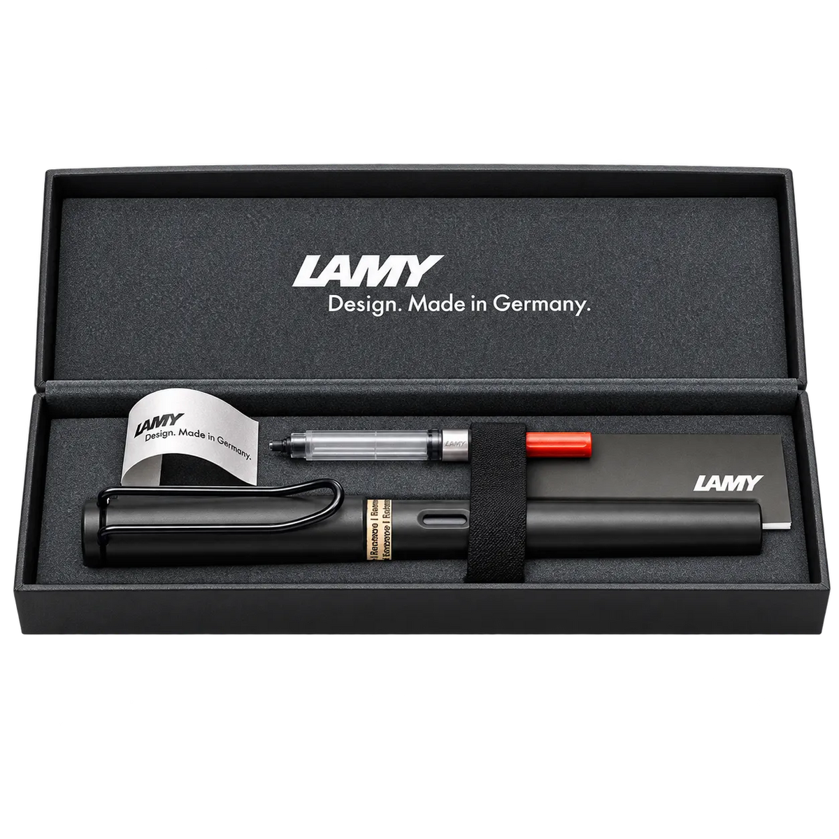 LAMY pen set in an open black presentation box, including a black pen, a clear and silver top-load pen, a small orange pen cap, and branded paper inserts on a white background.
